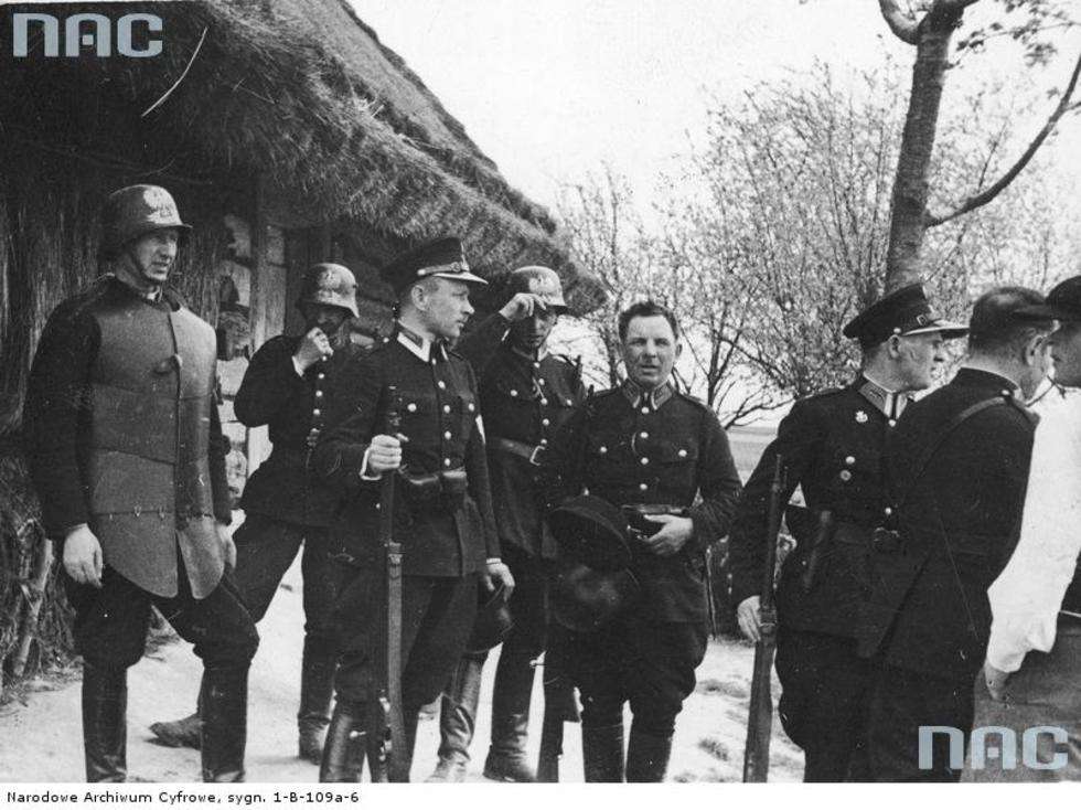  Funkcjonariusze policji przed wejściem do kryjówki bandy Maczugi. Z lewej widoczny policjant w kuloodpornej zbroi płytowej i hełmie, który miał wejść pierwszy do domu bandytów.

Data wydarzenia: 1934 
