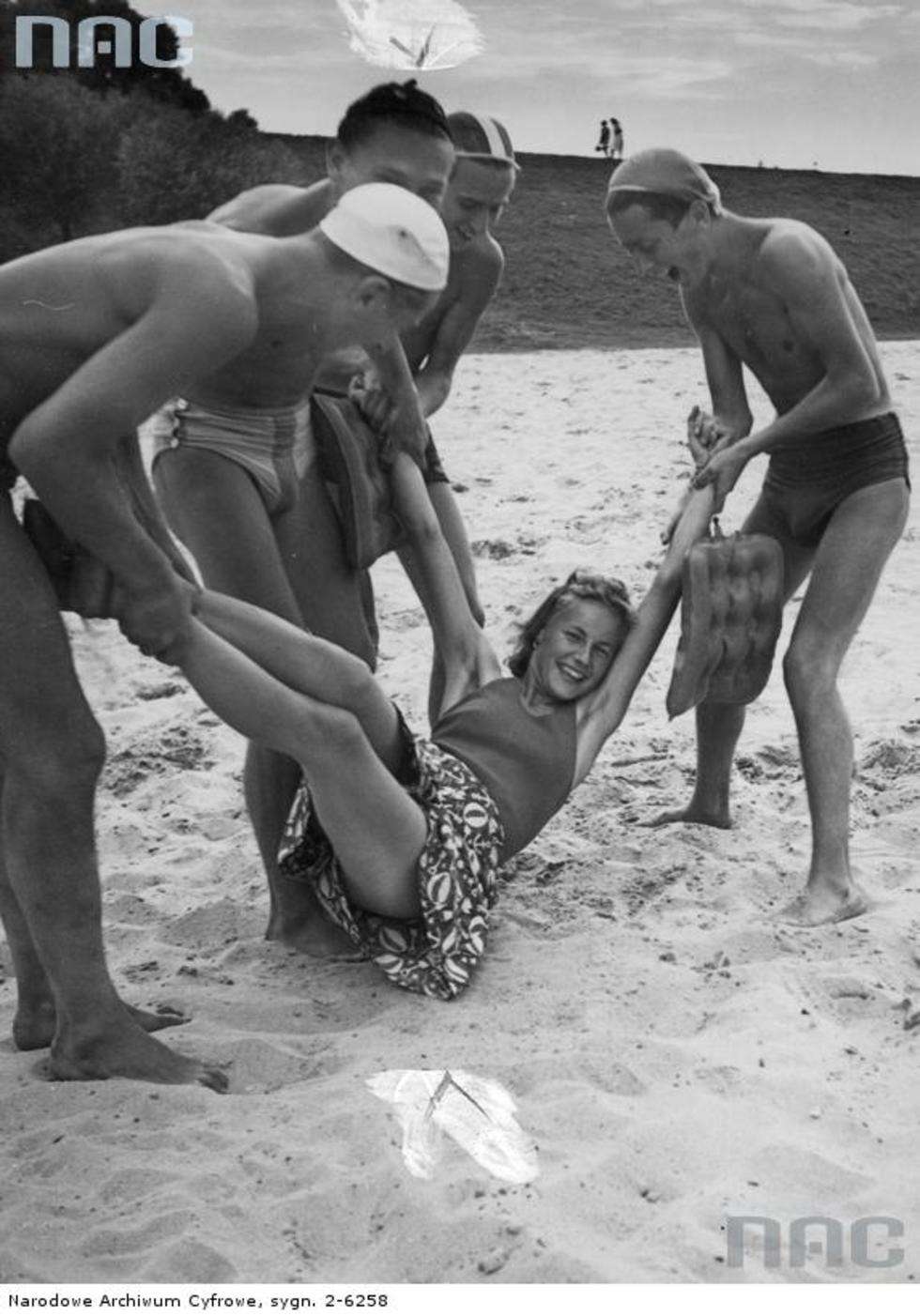 Kraków 1940 rok.  Pięć osób podczas zabawy na plaży nad Wisłą. Młodzi mężczyźni trzymają za ręce i nogi młodą kobietę. Fot. <a href="http://img.audiovis.nac.gov.pl/PIC/PIC_2-6258.jpg">NAC</a>