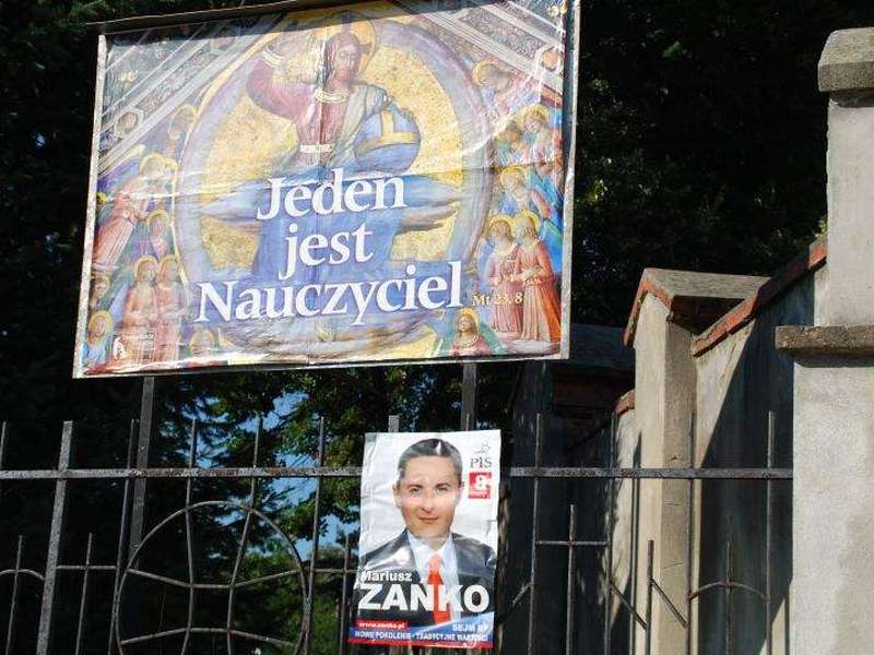 Plakat kandydata PiS obok tablicy z wizerunkiem Chrystusa i napisem "Jeden jest nauczyciel" (Joann