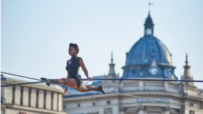 Kobieta wykonuje szpagat na rozpiętej w powietrzu linie. W tle dach Grand Hotelu w Lublinie